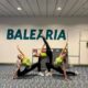 Tres gimnastas con camiseta verde posan en posturas de flexibilidad junto al letrero de Baleària