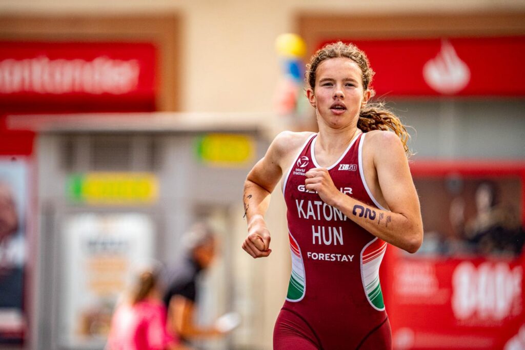 Young female athlete in a maroon racing suit runs in an urban street race, jersey reads 'KATONA HUN' with sponsor logos on the chest.