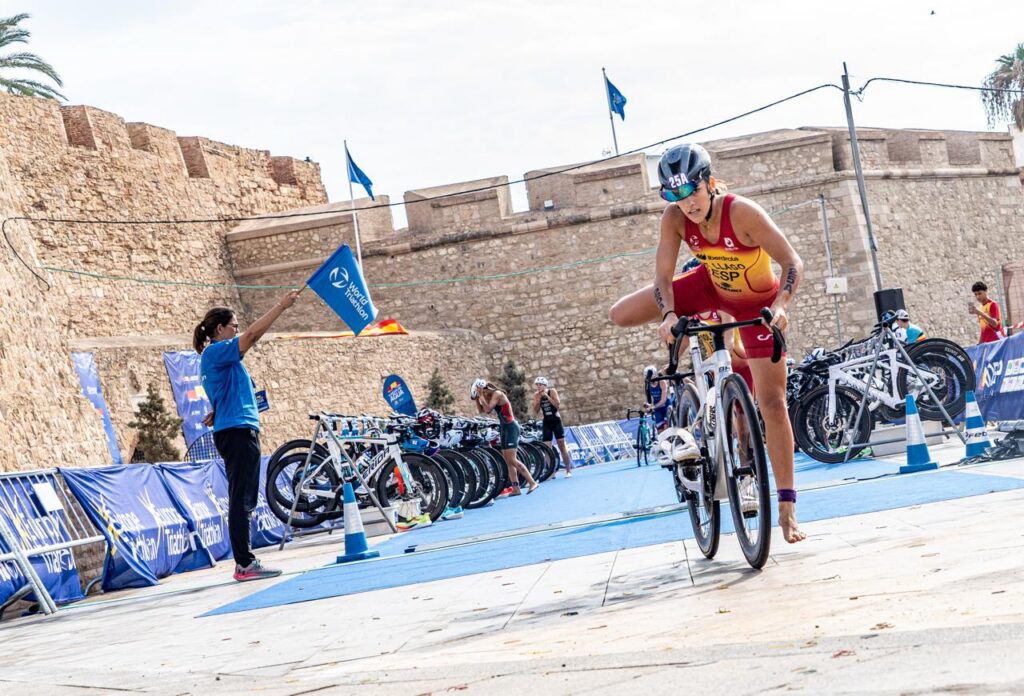 Una deportista con equipación ESP corre descalza junto a su bicicleta por la zona de transición.