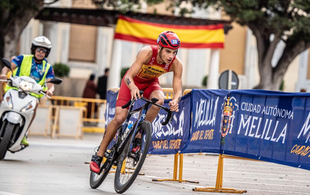 Un triatleta con mono rojo y amarillo pedalea entre vallas en una calle de Melilla
