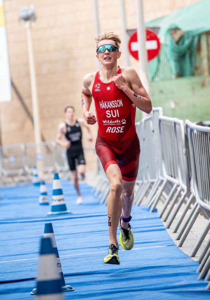 Un triatleta con uniforme suizo corre por una alfombra azul junto a vallas.