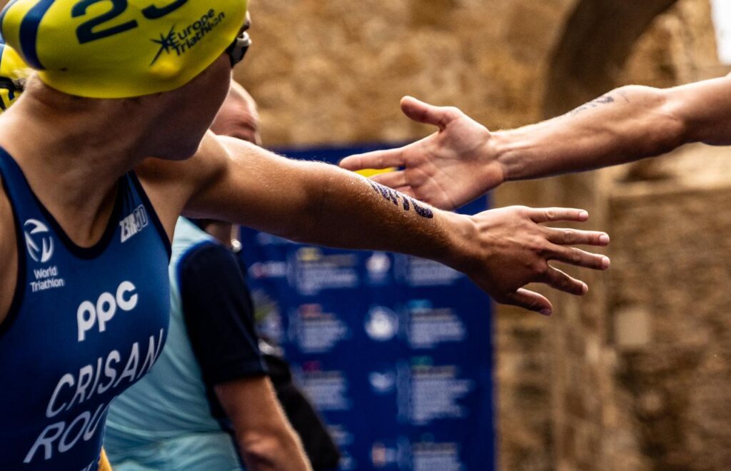 Dos triatletas extienden los brazos durante un relevo, con gorro amarillo y uniforme azul.