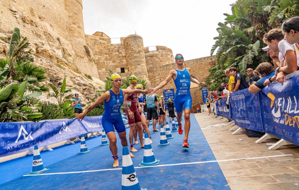 Triatletas realizan un relevo junto a espectadores, sobre una alfombra azul frente a murallas de piedra.