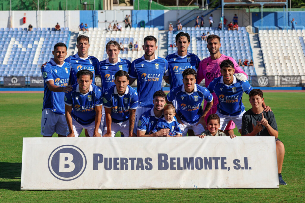 Jugadores de la U.D. Melilla posan con niños detrás de un cartel de patrocinio