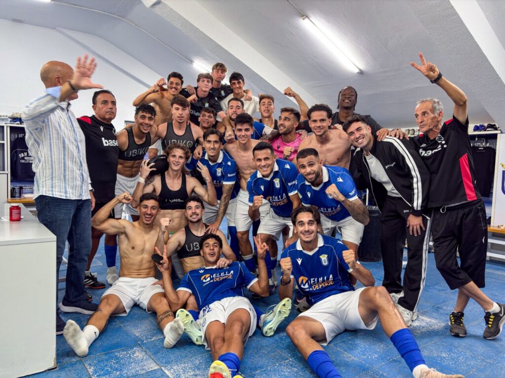Jugadores de fútbol posan juntos en un vestuario, varios levantan los brazos.