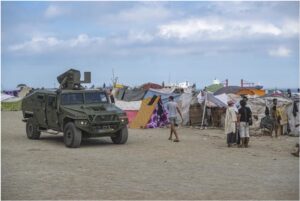 Vehículo militar junto a varias tiendas improvisadas y personas en un terreno abierto.