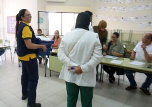 Volunteer in a yellow-and-navy uniform carries a blue box toward a registration desk where people sit with papers nearby.