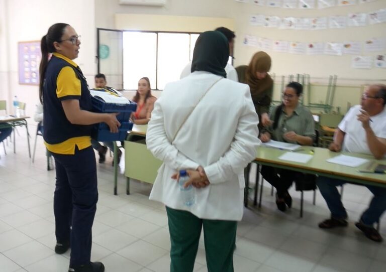 Volunteer in a yellow-and-navy uniform carries a blue box toward a registration desk where people sit with papers nearby.