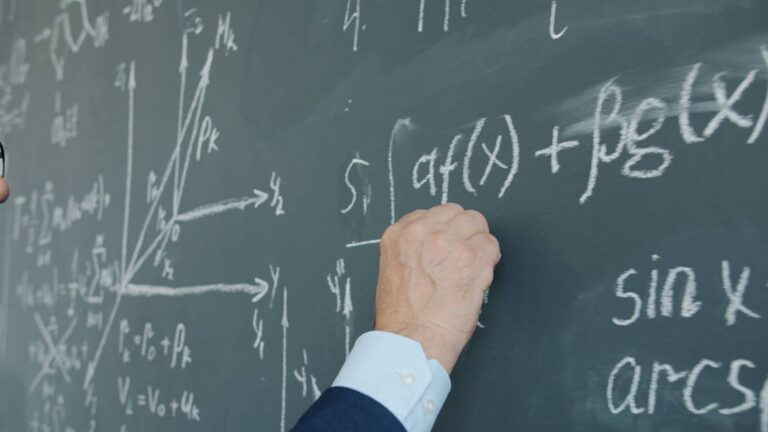 Una mano escribe fórmulas matemáticas con tiza en una pizarra verde.